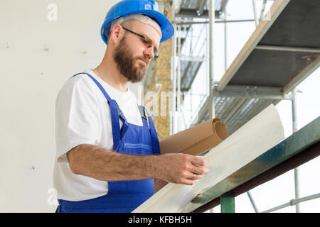 Construction worker in working outfit and in helmet stands at high altitude on construction site and inspects technical plans Stock Photo