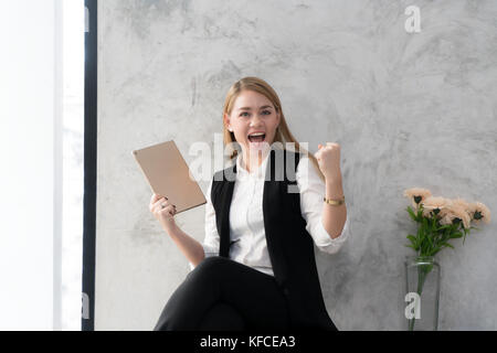 Happy businesswoman with raised in yes gesture hand reading tablet at desk in office. Female entrepreneur rejoicing good news from correspondence. Exi Stock Photo