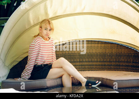 A young blond teenager girl in the street. Girl sitting on rottan sofa under the awning in restaurant Stock Photo