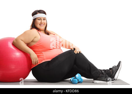 Overweight woman sitting on an exercise mat and leaning on a pilates ball isolated on white background Stock Photo