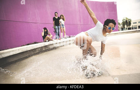 A young woman crouching down on a skateboard to create a water spray. Stock Photo