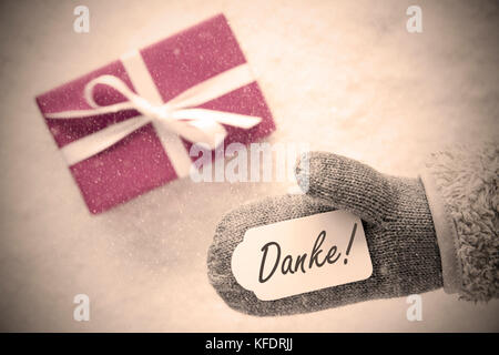 Glove With Label With German Text Danke Means Thank You. Pink Or Rose Gift Or Present On Snow In Background. Seasonal Greeting Card With Snowflakes An Stock Photo
