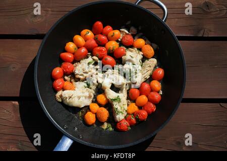fish with tomatoes and herbs on black pan standing on wooden planks Stock Photo
