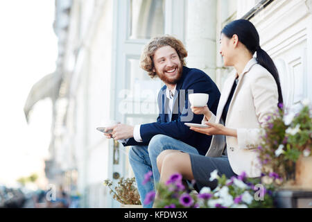 Young friendly colleagues with cups of coffee having talk at break in urban environment Stock Photo