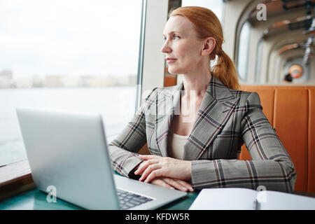 Waist-up portrait of attractive middle-aged businesswoman looking out window of middle deck while working on promising project with help of laptop Stock Photo
