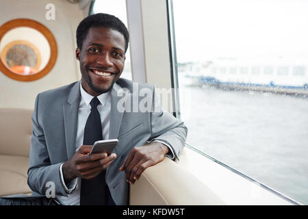 Waist-up portrait of cheerful African American white collar worker posing for photography while sitting at middle deck and browsing Internet on smartp Stock Photo