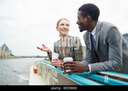 Cheerful coworkers chatting animatedly with each other and warming themselves with hot tea while standing on upper deck of ship Stock Photo