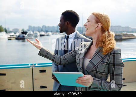 Profile view of attractive middle-aged businesswoman pointing at something while standing on upper deck with African American colleague and discussing Stock Photo