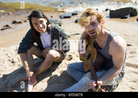 Young man playing guitar and singing on seashore with his friend on background Stock Photo