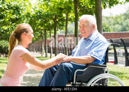 Happy Young Woman Holding Hands Of Her Disabled Father On Wheelchair In Park Stock Photo