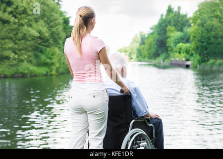 Rear View Of A Young Woman With Her Disabled Father On Wheelchair Looking At Lake Stock Photo