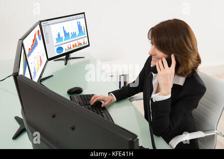 Businesswoman Analyzing Graphs On Multiple Computers While Talking On Phone Stock Photo