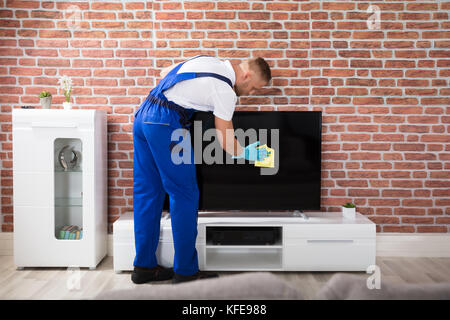 Male Janitor In Uniform Cleaning The Television With Rag Stock Photo