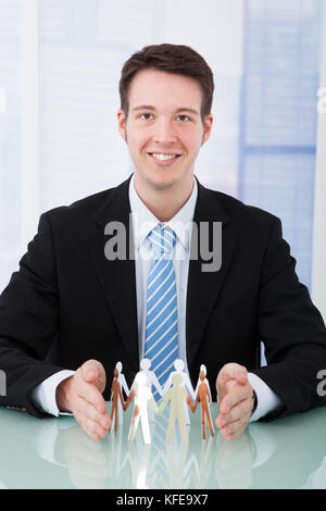 Portrait of confident young businessman protecting team of paper people at office desk Stock Photo