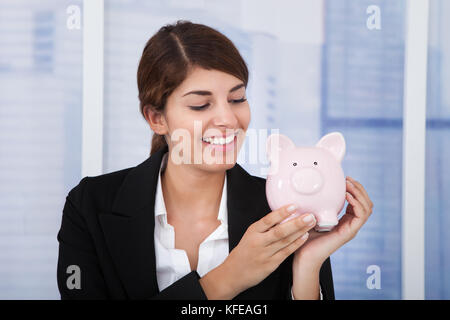 Portrait of happy young businesswoman holding piggybank in office Stock Photo