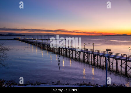 Wooden pier at White Rock, BC, Canada extends diagonally into image... its twinkling lights reflected in the sea.  Setting sun with deep colour. Stock Photo
