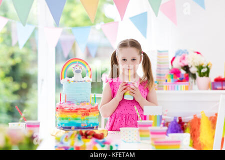 Kids birthday party with colorful pastel decoration and unicorn rainbow cake. Little girl with sweets, candy and fruit. Balloons and banner at festive Stock Photo