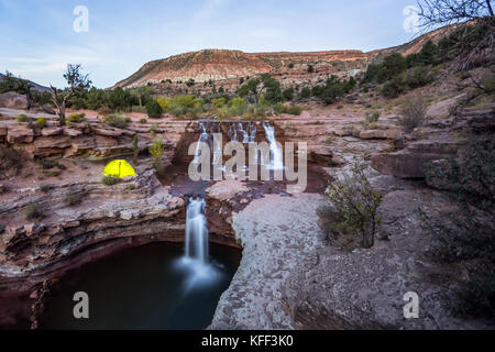 A yellow-green tent lit up from the inside rests on a rocky cliff near a cascade of several waterfalls leading to a larger fall below. After sunset, a Stock Photo