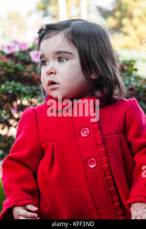 Cute, pretty, happy and fashionable toddler baby girl, wearing a good fashion red long coat or overcoat on cold winter day in a garden. 18 months old Stock Photo