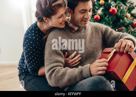 Young man opening Christmas gift. Smiling woman holding her partner from behind while spending happy moments celebrating Christmas. Stock Photo