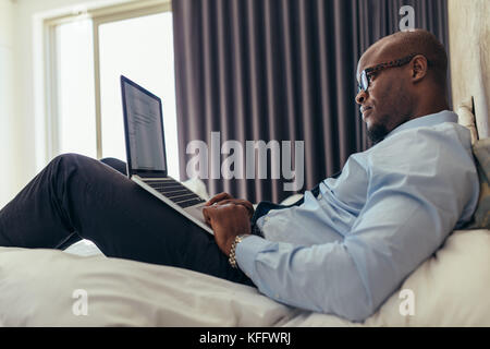 Man in formal clothes working on laptop while lying in bed. Businessman working on laptop computer from his hotel room. Stock Photo