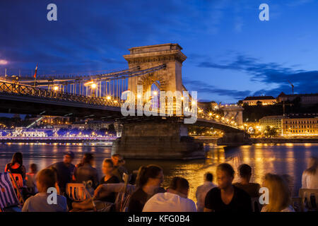 Budapest by night in Hungary, city break, group of people hangout and chill out at Danube river next to Szechenyi Chain Bridge, both local and tourist Stock Photo