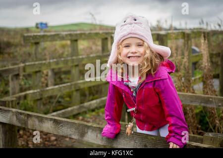 Young female child with a big smile dressed in autumn winter clothes enjoying the outdoors on a walk in the countryside. She is climbing on a fence. Stock Photo