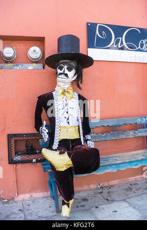 jaunty life size Day of the Dead gentleman with repaired plaster head hands & chipped top hat sits outside shop in tacky velvet & lace formal attire Stock Photo