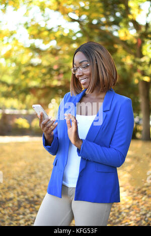 Black woman in blue blazer standing outdoors while using her smartphone. Stock Photo