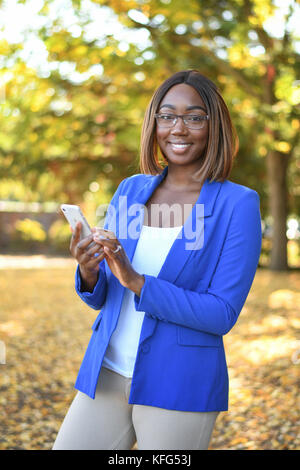 Black woman in blue blazer standing outdoors while using her smartphone. Stock Photo