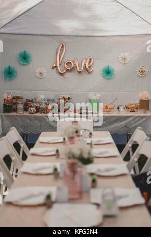 Long white table setting with white chairs lined up. Background shows assorted candy in jars on food table with shower party decor and "love" sign Stock Photo