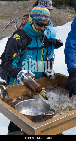 Local tour guide poring small glasses of whiskey over glacier ice at end of hiking on glacier.  Bottle in hand with ice ax strapped on back. Stock Photo