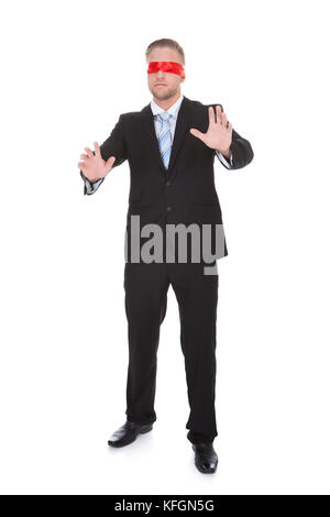 Stylish young businessman wearing a red blindfold stretching his hands out in front of him to feel for unseen obstacles  full length isolated on white Stock Photo
