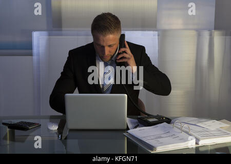 Stressed businessman working late into the night putting in overtime to meet a deadline sitting at his desk using a laptop and talking on his mobile Stock Photo
