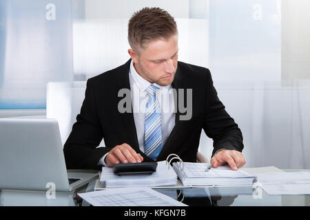 Hardworking businessman analyzing a report sitting at his desk using a calculator and reading a report in a binder