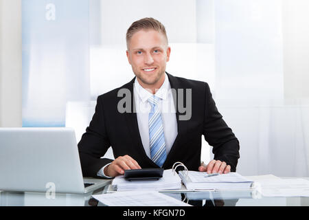 Smiling successful businessman sitting st his desk in the office surrounded by paperwork using a calculator and a laptop