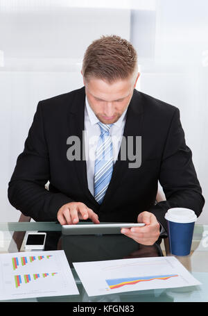 Stylish hardworking businessman using a tablet computer at his desk concentrating as he works online with graphs spread out in front of him Stock Photo