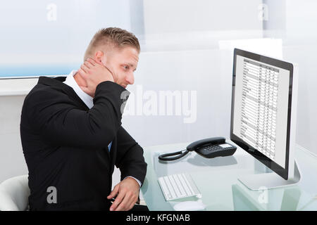 Businessman studying an online spreadsheet on a desktop monitor rubbing his neck in confusion or to ease stiffness Stock Photo