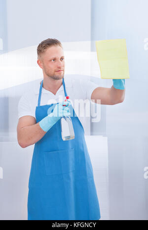 Friendly smiling male cleaner cleaning a pane of glass with a spray bottle and cloth in an office building Stock Photo