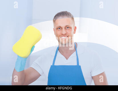 Male janitor using a sponge to clean a window in an office wearing an apron and gloves as he works Stock Photo