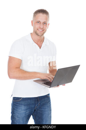 Smiling man in jeans standing using a handheld laptop computer balanced on his palm as he looks at the camera with a friendly smile  isolated on white Stock Photo