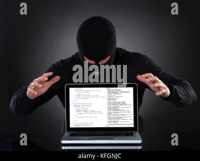Hacker in a balaclava standing in the darkness furtively stealing data off a laptop computer or inserting spyware in an online security and risk conce Stock Photo