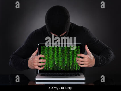 Hacker in a balaclava standing in the darkness furtively stealing data off a laptop computer or inserting spyware in an online security and risk conce Stock Photo