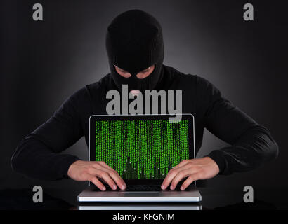 Hacker in a balaclava standing in the darkness furtively stealing data off a laptop computer or inserting spyware in an online security and risk conce Stock Photo