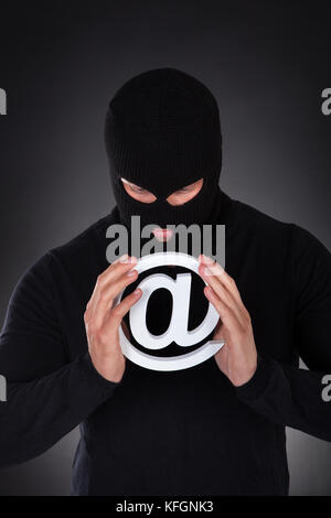 Hacker dressed in black wearing a balaclava with a white internet domain symbol in his hands as he stands in darkness conceptual of online safety and  Stock Photo