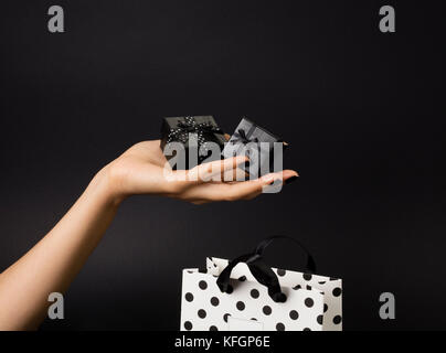 Female hand holding a three black gift-boxes on a palm, black gift-bag decorated with white polka dots below. Black background. Stock Photo