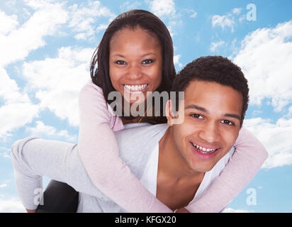 Portrait of happy young man giving piggyback ride to woman against sky Stock Photo