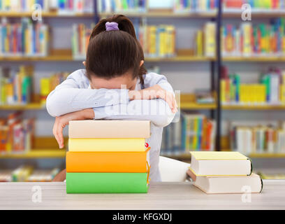 Tired female college student leaning on stack of books at desk in library Stock Photo