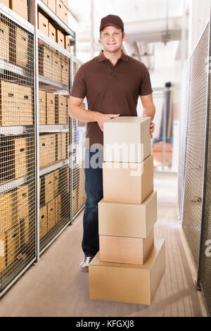 Portrait of smiling delivery man with stacked cardboard boxes standing in warehouse Stock Photo
