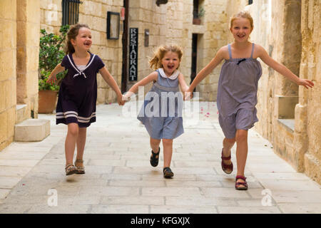 Three young sisters / girls / children / kids / kid aged 7, 3, & 5 years old, on a family holiday, run & play and span a narrow street in Mdina, Malta